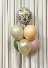 Load image into Gallery viewer, A festive Easter balloon bouquet featuring a large foil balloon with &#39;Happy Easter&#39; written in gold lettering, surrounded by illustrations of bunnies, eggs, and flowers. The foil balloon is complemented by pastel latex balloons in shades of cream, peach, sage green, and blush pink, tied together with ribbons against a white curtain backdrop
