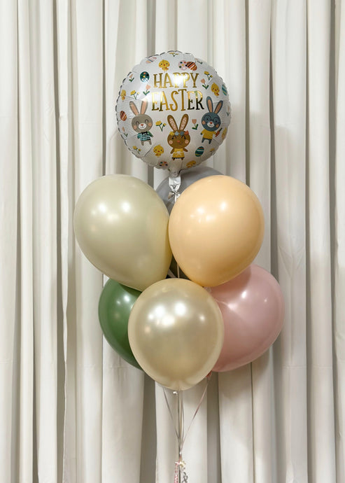 A festive Easter balloon bouquet featuring a large foil balloon with 'Happy Easter' written in gold lettering, surrounded by illustrations of bunnies, eggs, and flowers. The foil balloon is complemented by pastel latex balloons in shades of cream, peach, sage green, and blush pink, tied together with ribbons against a white curtain backdrop