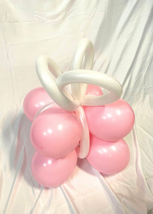 A festive balloon creation shaped like a gift box with pink balloons forming the base and white twisted balloons creating a ribbon bow on top