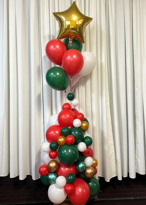 A detailed Christmas Tree Balloon Column topped with a shiny, gold foil star balloon. This arrangement features a mix of red, green, and white balloons in different sizes to form a tree shape. The balloons are layered, with larger balloons at the bottom and smaller ones leading up to the gold star at the top, creating a visually striking holiday display. The column includes metallic accents with some gold and green balloons, adding sparkle.