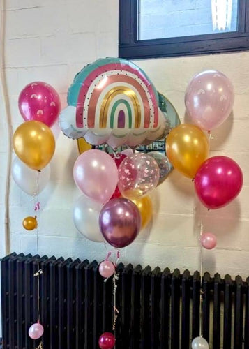 A bouquet of balloons featuring a large rainbow-shaped balloon, with a mix of round balloons in colors gold, pink, white, and magenta, along with heart-shaped balloons in pink.