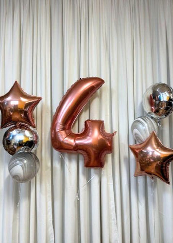 A set of birthday balloons featuring rose gold and silver colors, with stars and number shapes, displayed against a white curtain backdrop.