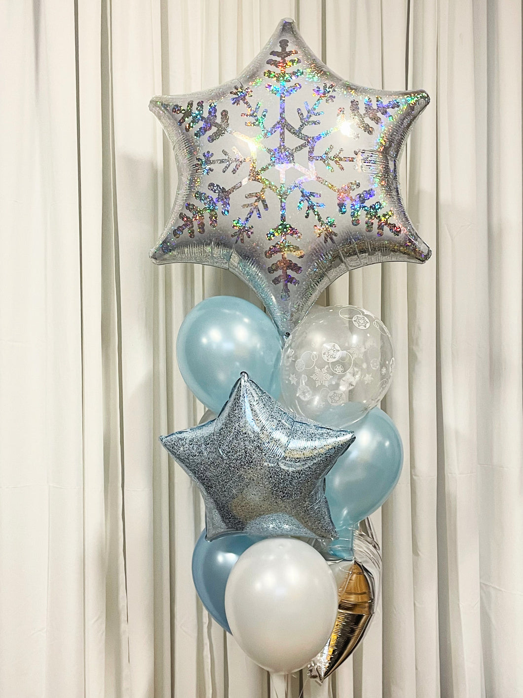 A bouquet of balloons featuring a large snowflake-shaped balloon, with smaller round balloons in blue and white shades, and decorative star-shaped balloons in silver and blue.